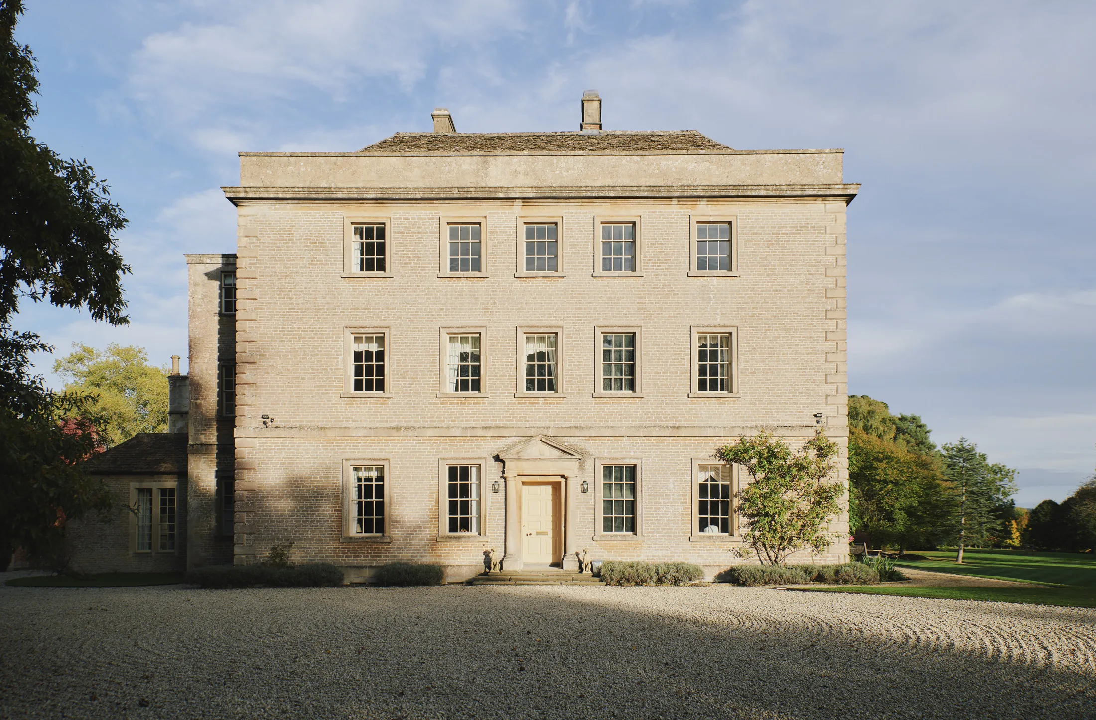 Elegant three-story stone manor with symmetrical windows and tree-lined gravel drive at Havenwood in The Cotswolds, a luxury short stay retreat.