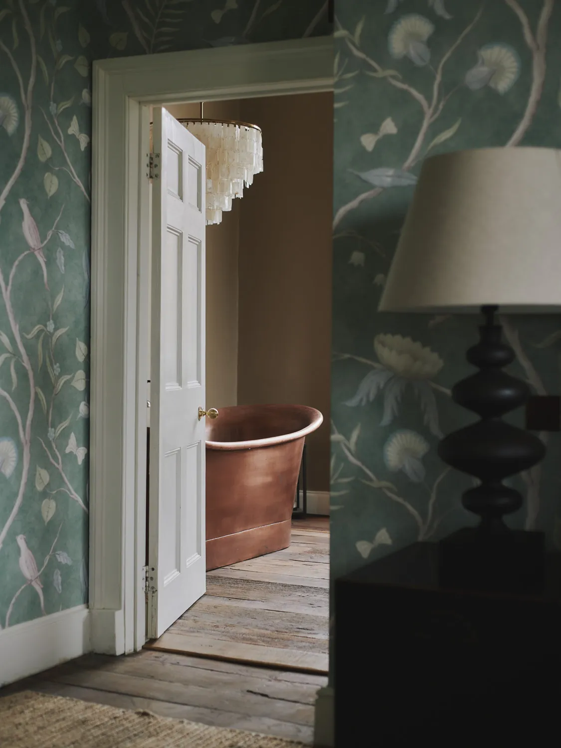 Freestanding copper bathtub glimpsed through doorway with shell chandelier at Havenwood exclusive countryside rental in The Cotswolds.