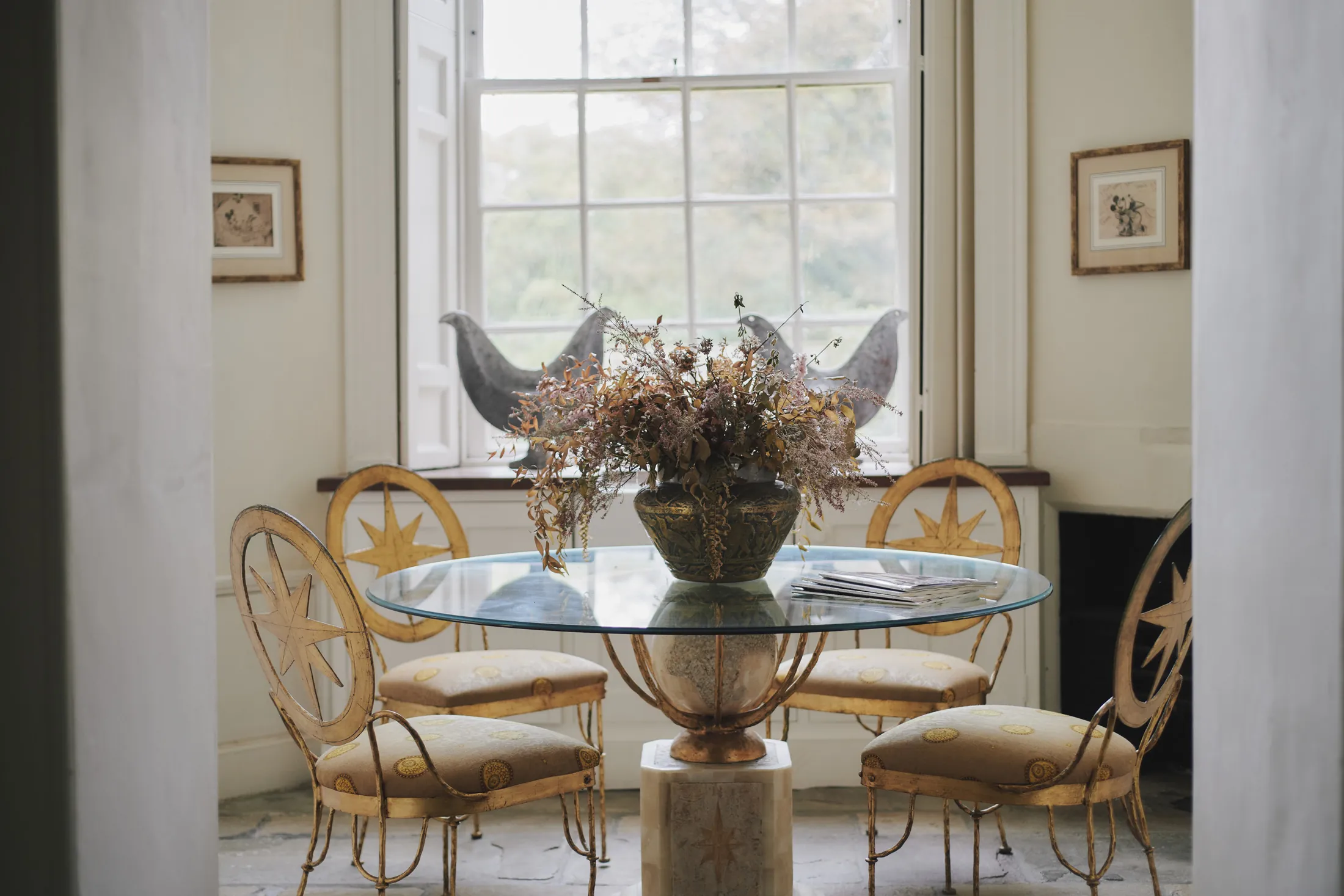 Gilded dining chairs surrounding glass table with fresh flowers near tall windows at Havenwood, a Domus Stay luxury getaway.