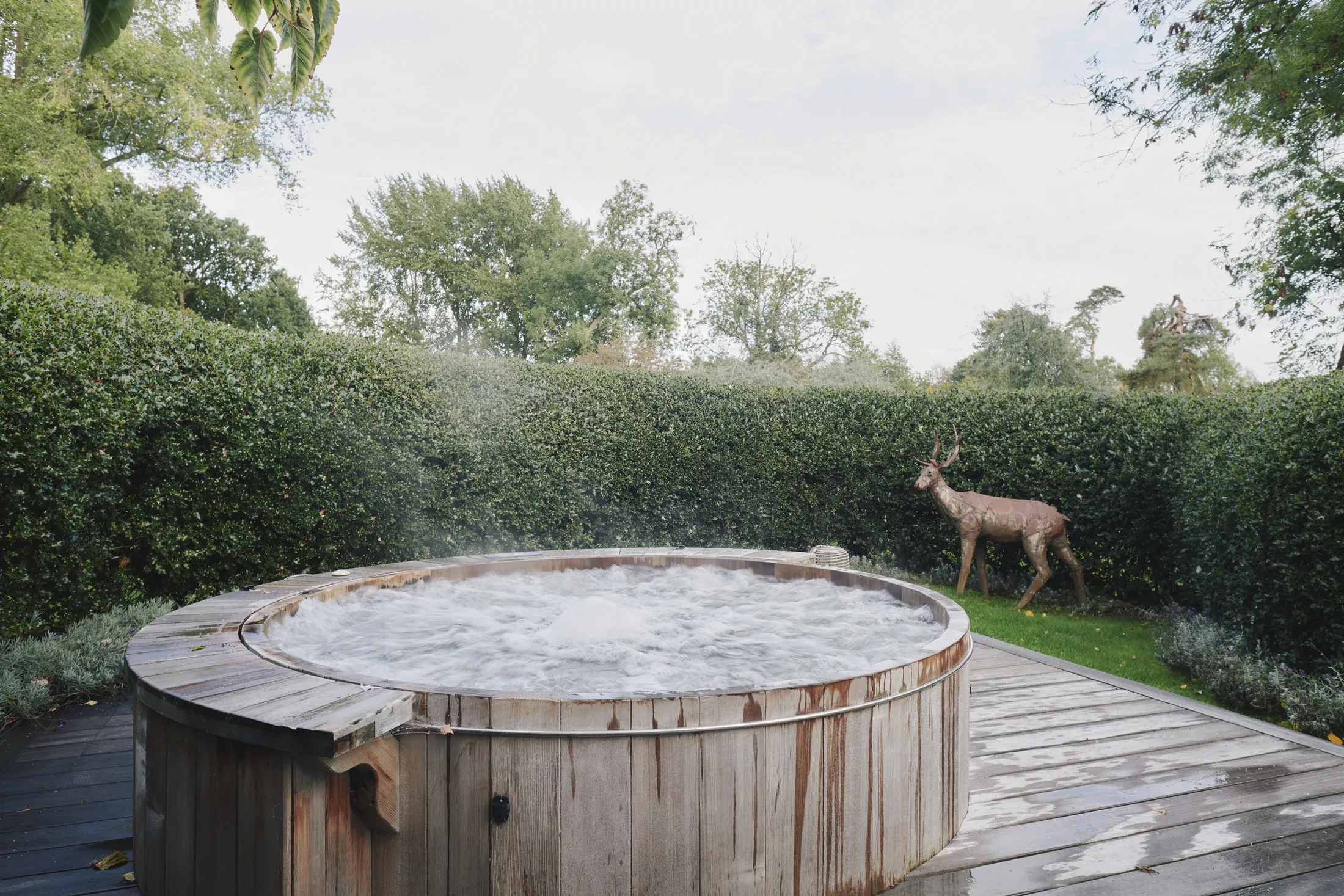 Wooden hot tub on decking surrounded by manicured hedges and garden sculpture at Havenwood, a Domus Stay luxury short stay in The Cotswolds.