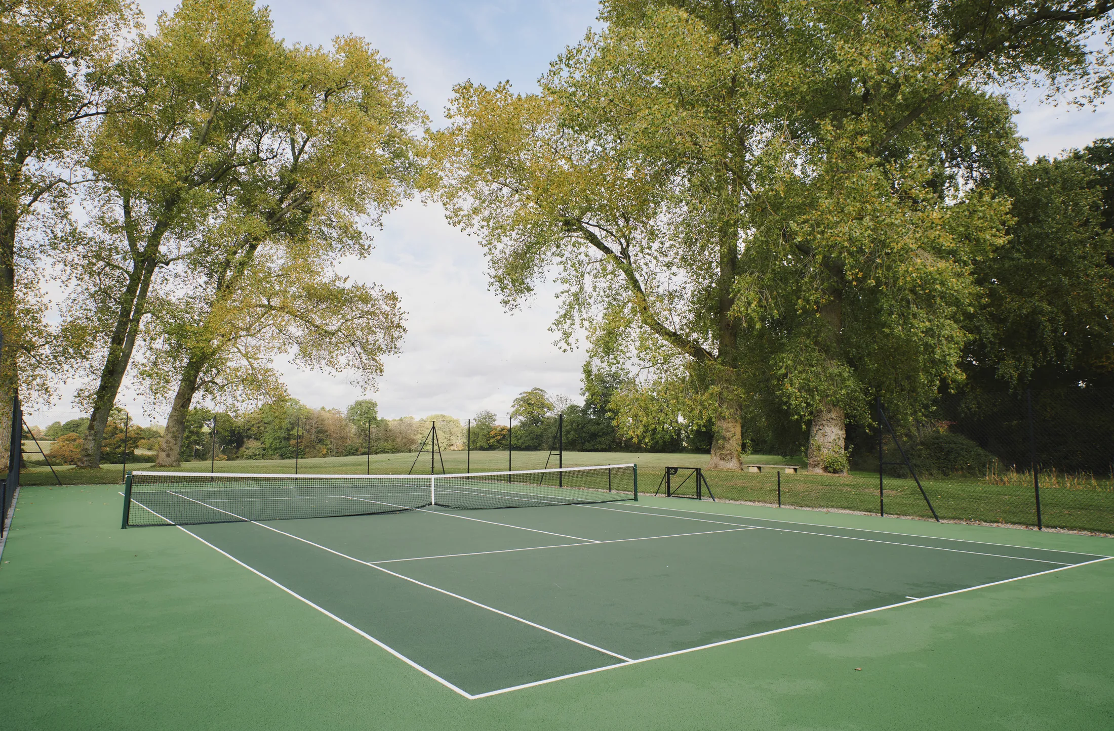 Tranquil tennis court under clear blue skies surrounded by lush greenery at Havenwood, a Domus Stay countryside retreat in The Cotswolds.