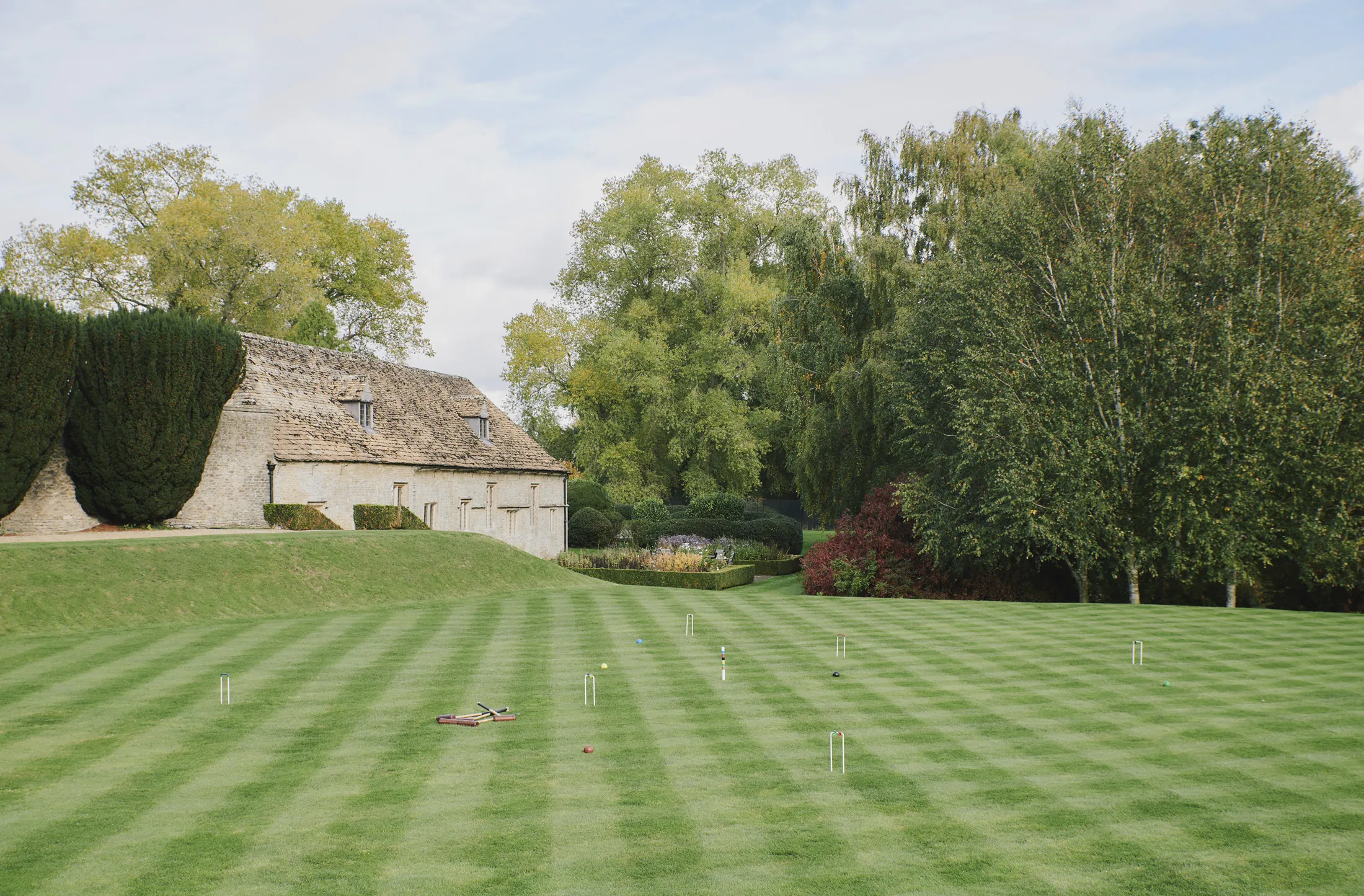 Stone cottage overlooking pristine croquet lawn framed by mature trees at Havenwood, The Cotswolds luxury countryside getaway.