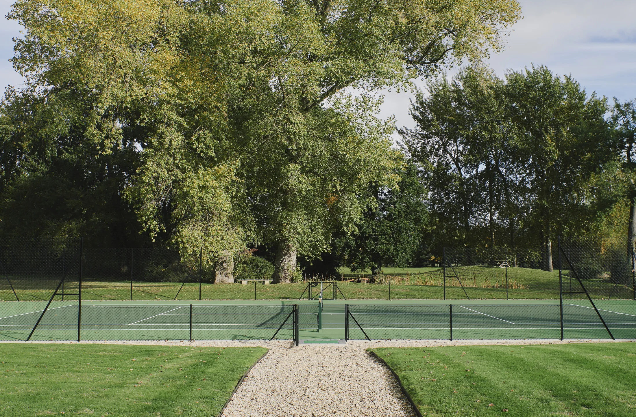 Private tennis court nestled among tall trees with gravel pathway at Havenwood, The Cotswolds luxury short term rental.