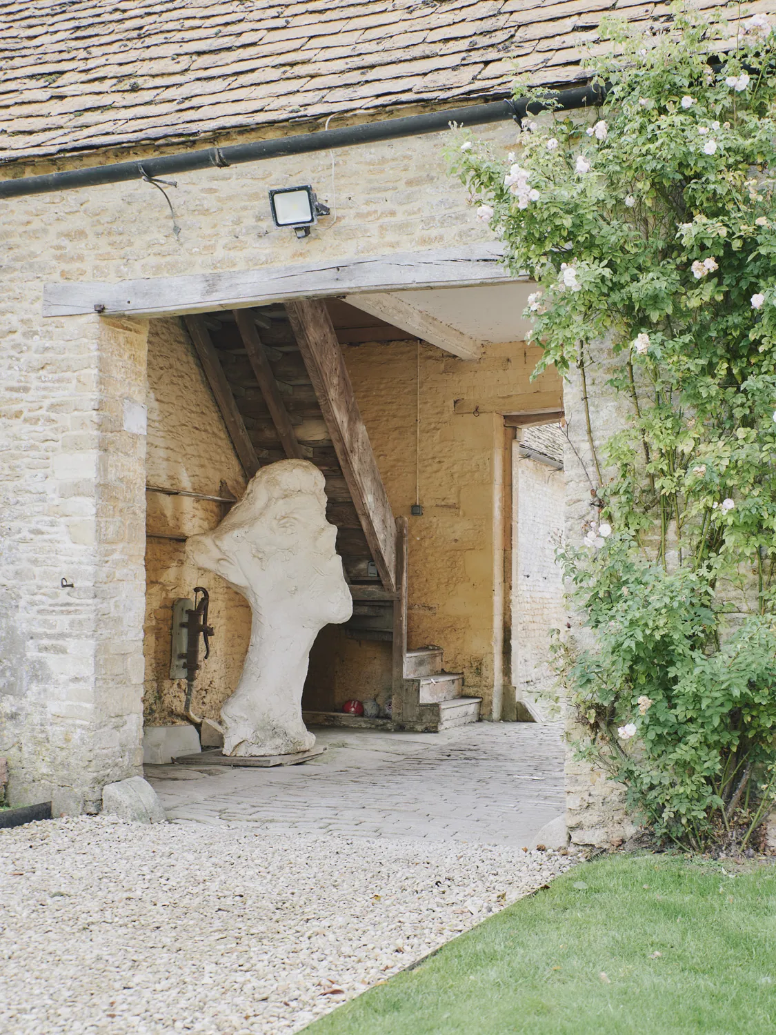 Sculptural stone head beneath exterior staircase adorned with climbing roses at Havenwood exclusive rental in The Cotswolds.
