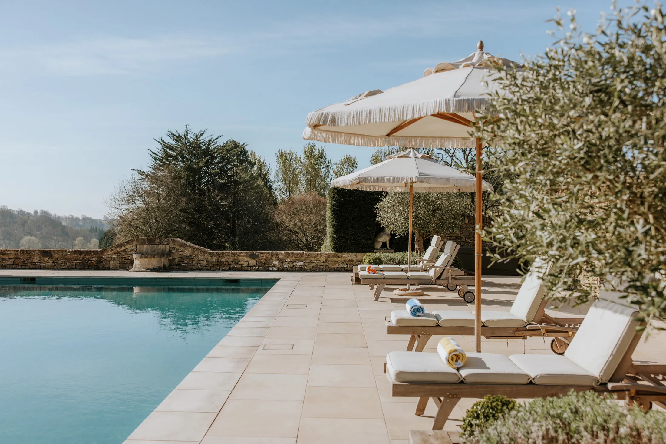 Sunlit poolside terrace at Coombe End, a Domus Stay luxury holiday rental, with cushioned loungers, parasols and views across the Cotswolds valley.