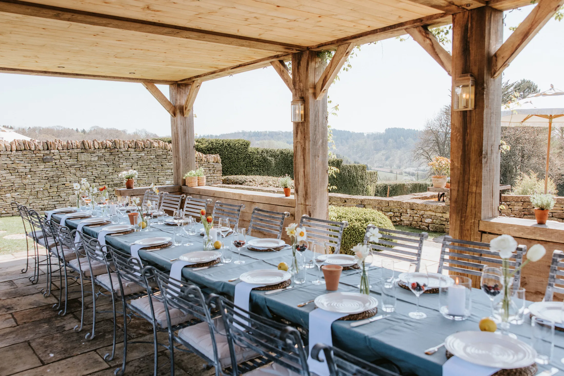 Covered outdoor dining terrace at Coombe End, part of the Domus Stay luxury rental portfolio, overlooking Cotswolds scenery with a long banqueting table.