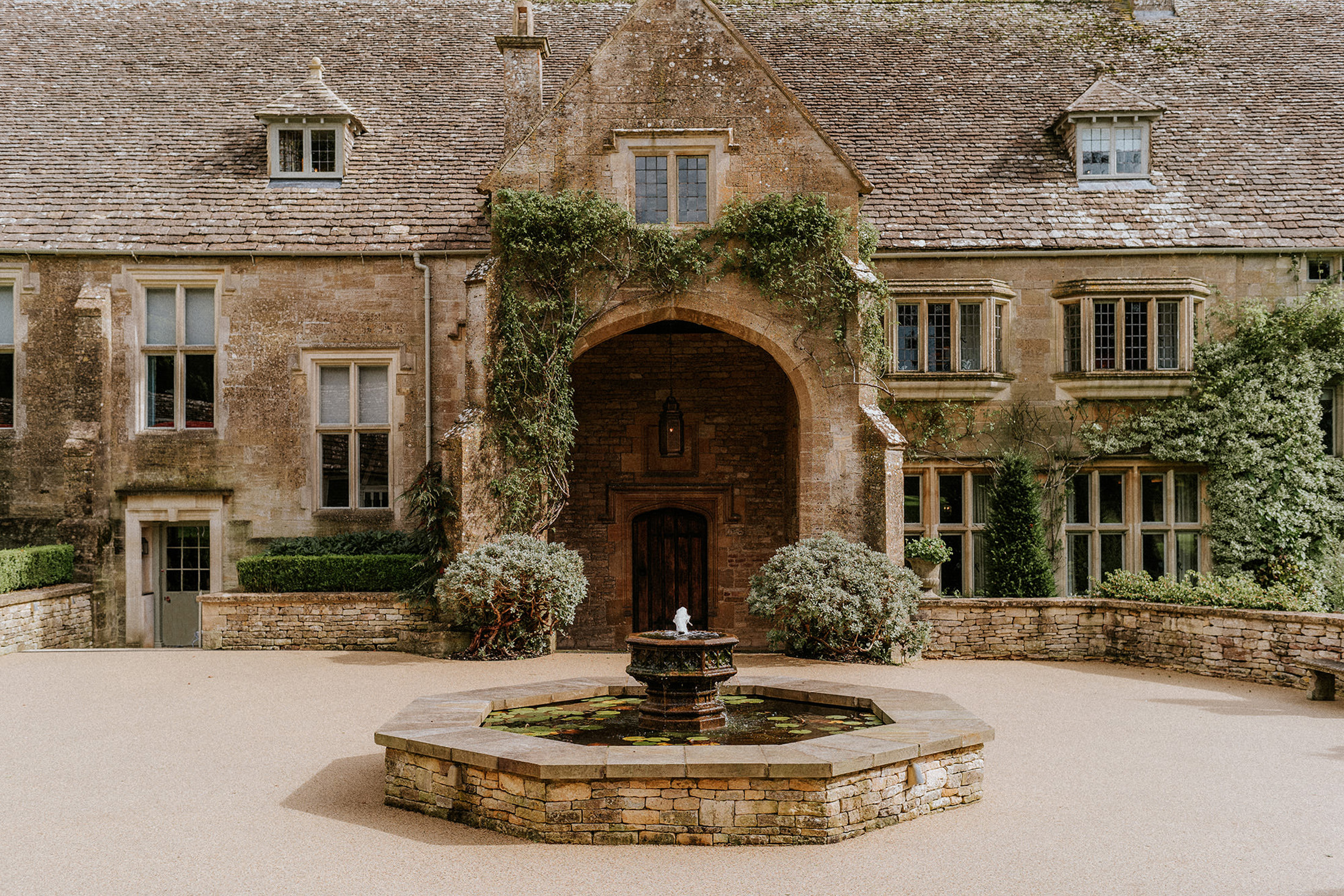 Grand Cotswolds manor entrance at Coombe End, a Domus Stay premium short-stay property, framed by climbing greenery and a central stone fountain.
