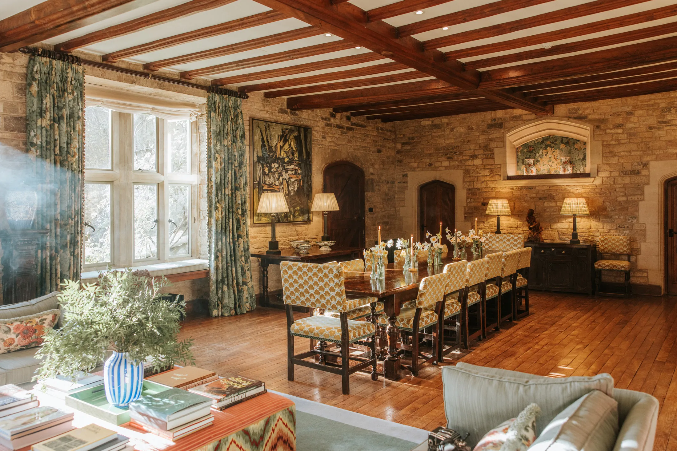 Warm dining hall at Coombe End, a Domus Stay countryside short-stay, showcasing a long oak table, patterned chairs and mullioned windows with floral drapes.