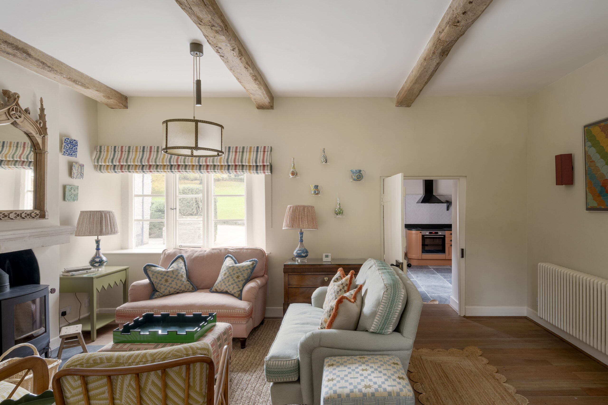 Charming sitting room at Ford Cottage, a Domus Stay luxury Cotswolds rental, with pastel sofas, striped blinds, eclectic wall art and views across the cottage garden.