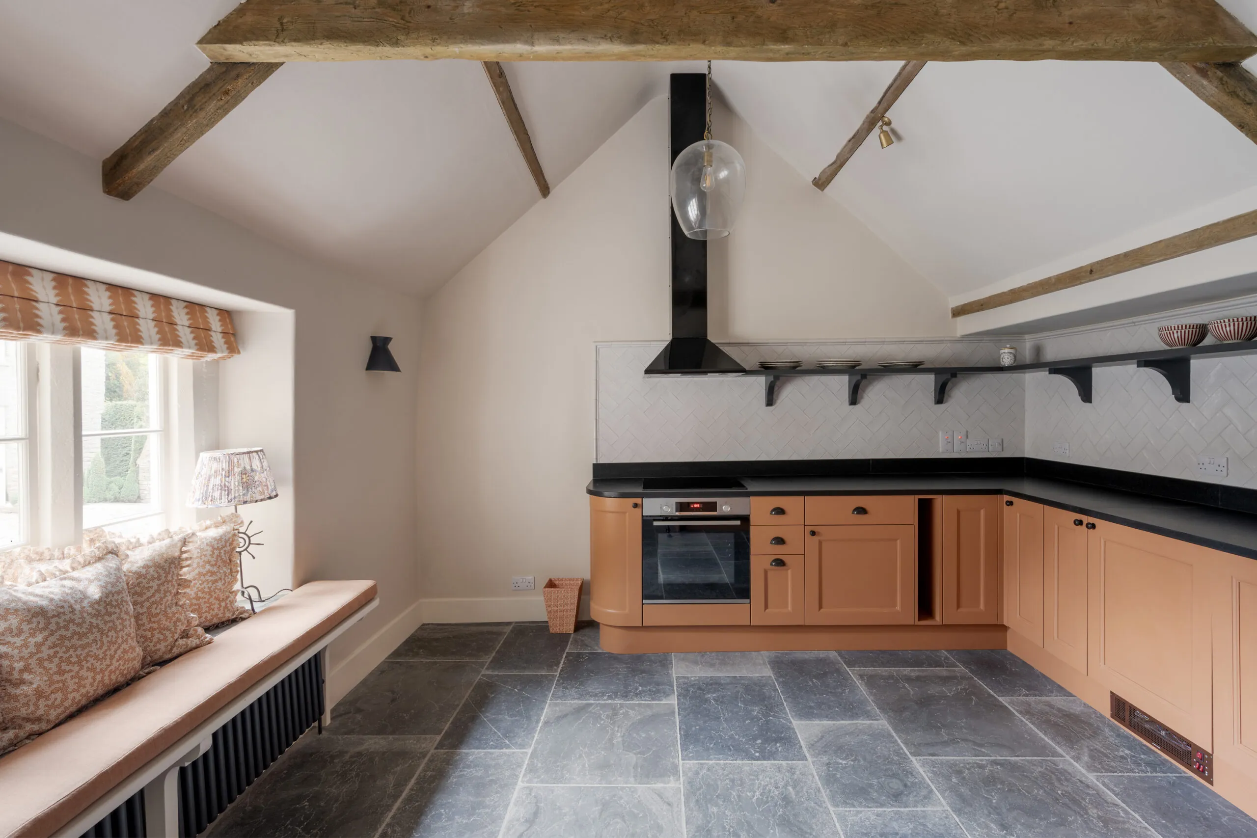 Beautiful country kitchen at Ford Cottage, offered by Domus Stay as a luxury short stay rental in The Cotswolds, with peach cabinetry, exposed beams, slate flooring and a deep garden-facing window seat.