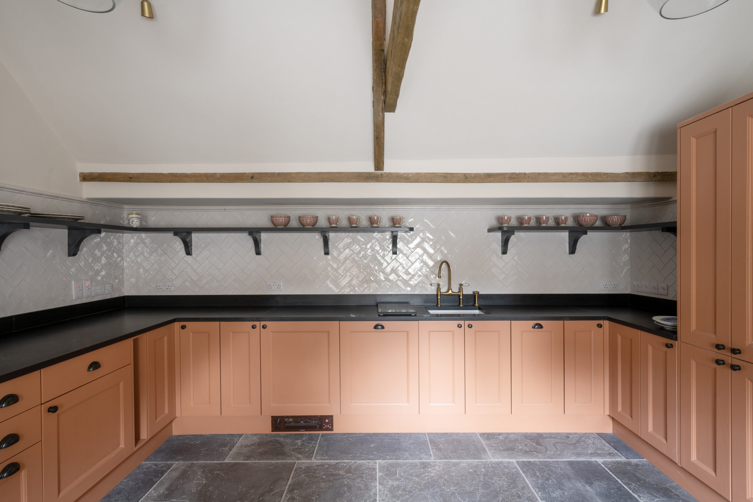 Peach-toned kitchen at Ford Cottage, a Domus Stay luxury short stay retreat in The Cotswolds, featuring herringbone tiles, brass taps, slate floors and open shelving with artisan bowls.